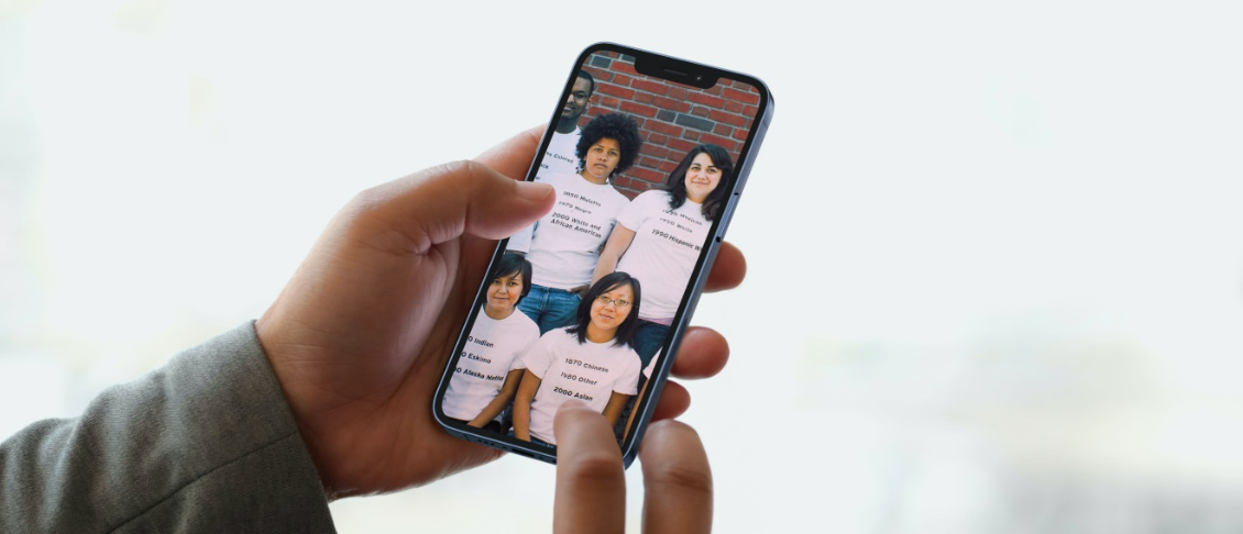 Two hands holding a smart phone. On the phone is an image of a group of individuals standing in a group, with historic census racial demographics on their shirts.