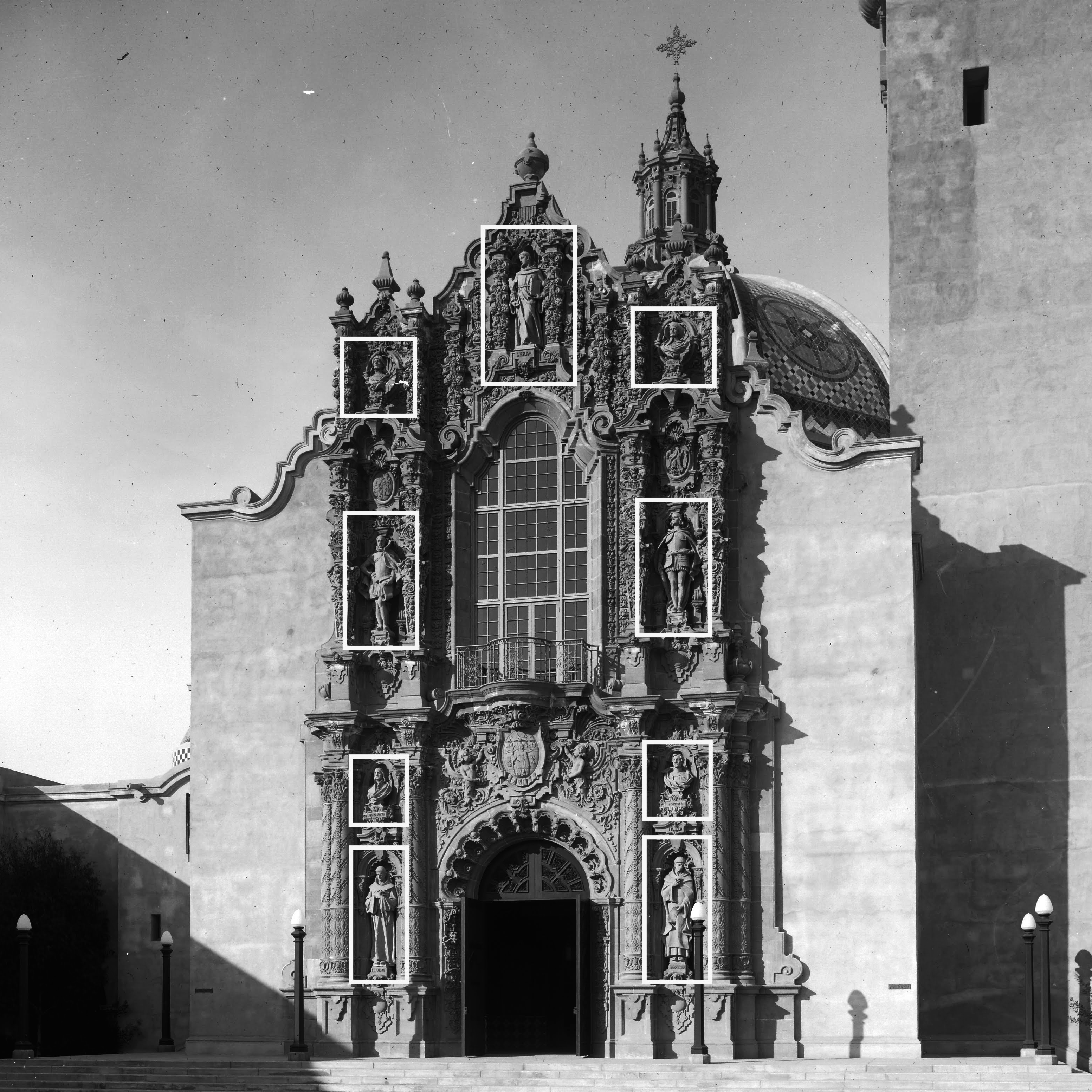 A vintage black and white photo of the Museum of Us façade. The nine colonizers etched into the Museum's exterior architecture are called out by white rectangles overlaid their statues.